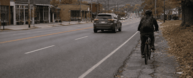 A person bicycling and a pedestrian use designated spaces alongside motor vehicles, illustrating the need for roadway safety strategies that protect every traveler.
